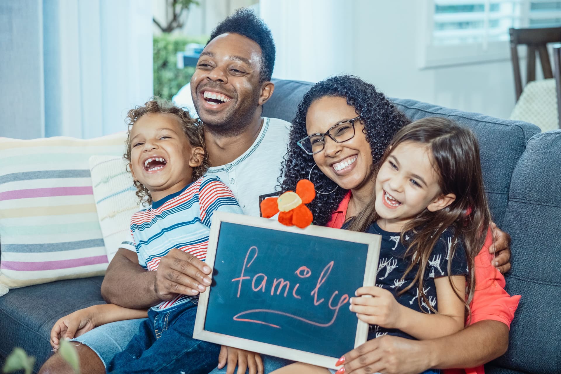 A smiling family sitting together on a couch holding a small chalkboard with the word family